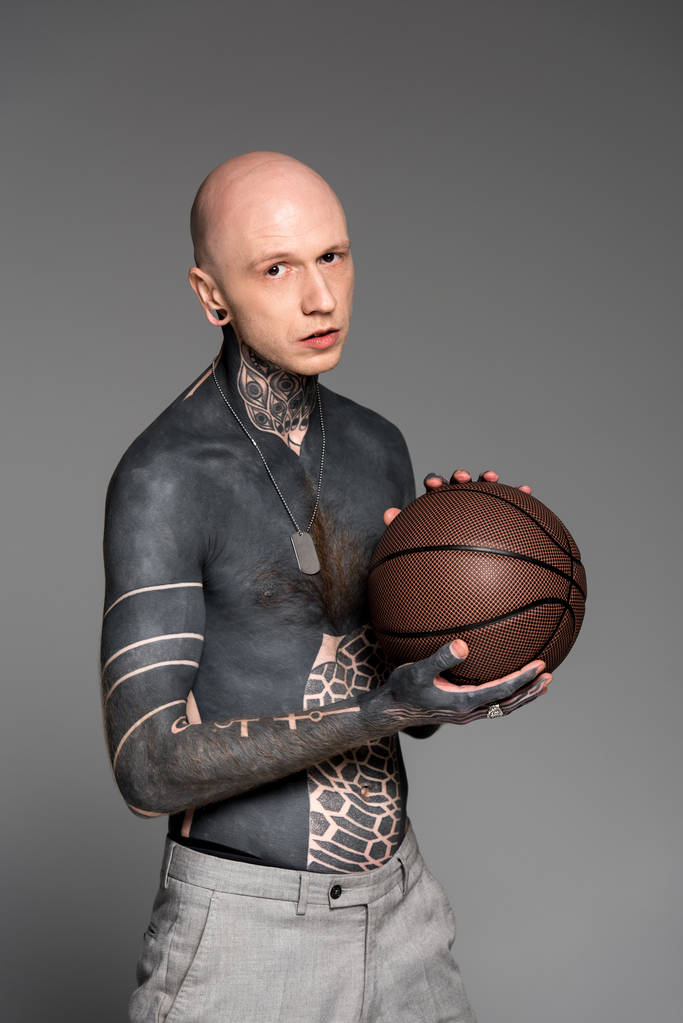 hombre de pecho desnudo con tatuajes sosteniendo pelota de baloncesto y mirando a la cámara aislada en gris
 - Foto, Imagen
