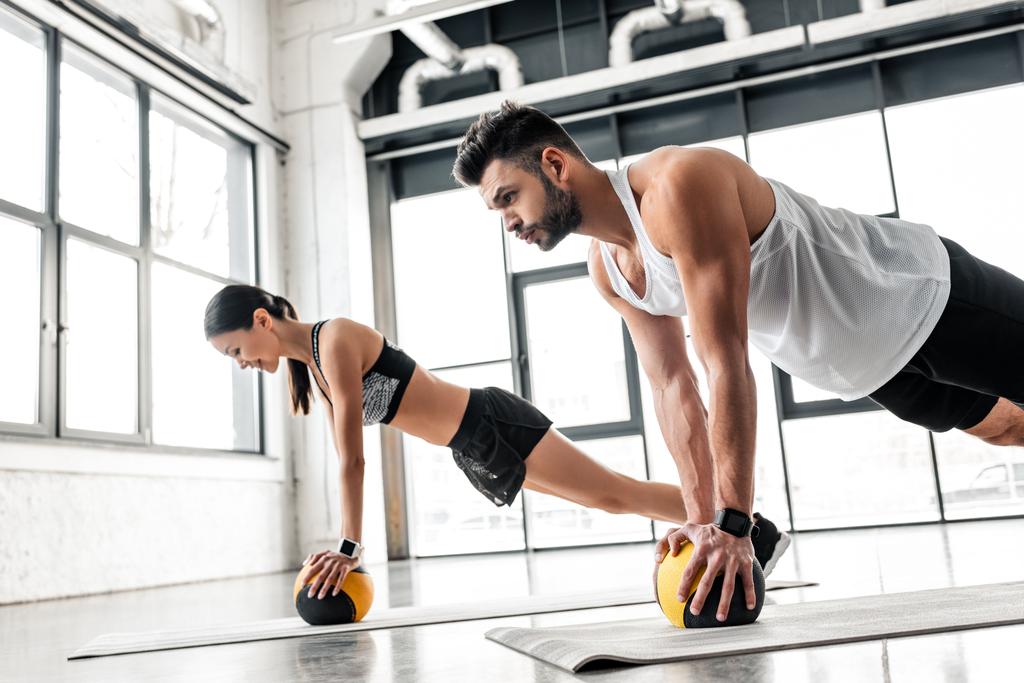 side view of athletic young couple in sportswear exercising with medicine balls on yoga mats in gym - Photo, Image