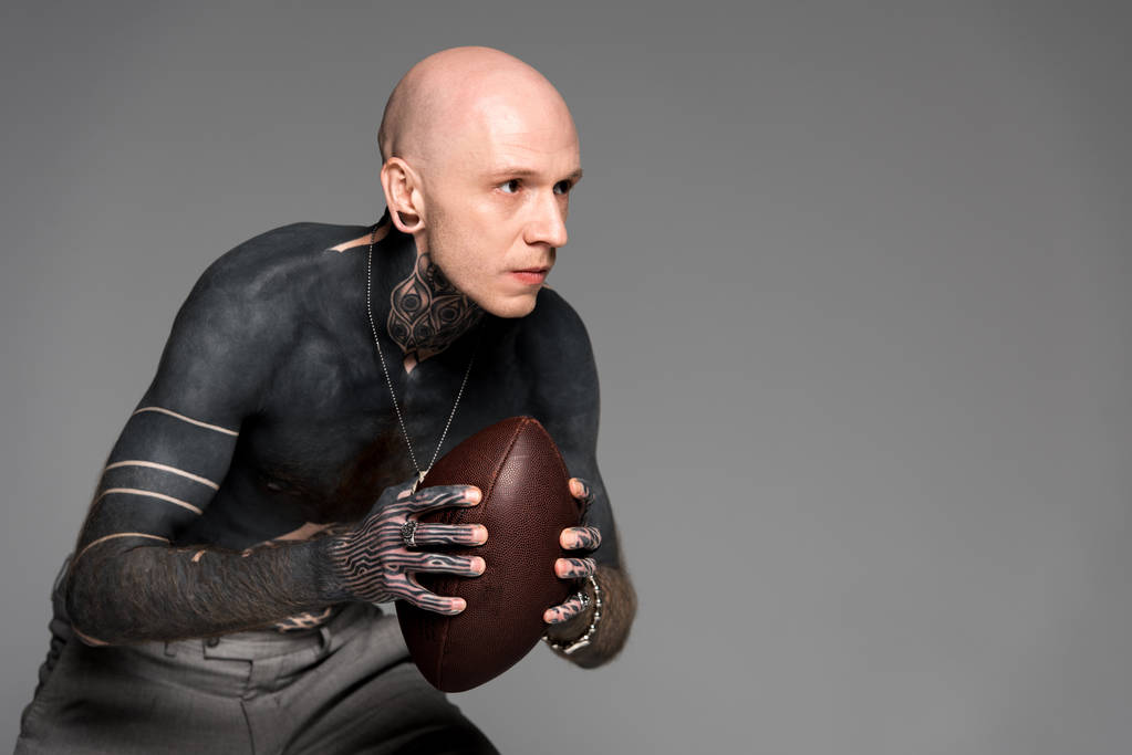 concentrated bare-chested tattooed man holding rugby ball and looking away isolated on grey  - Photo, Image