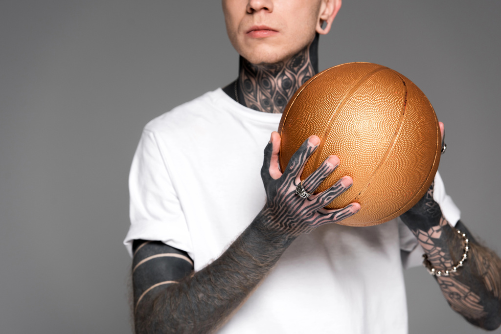 обрізаний знімок татуйованого чоловіка в білій футболці, що тримає золотий баскетбольний м'яч ізольований на сірому
 - Фото, зображення