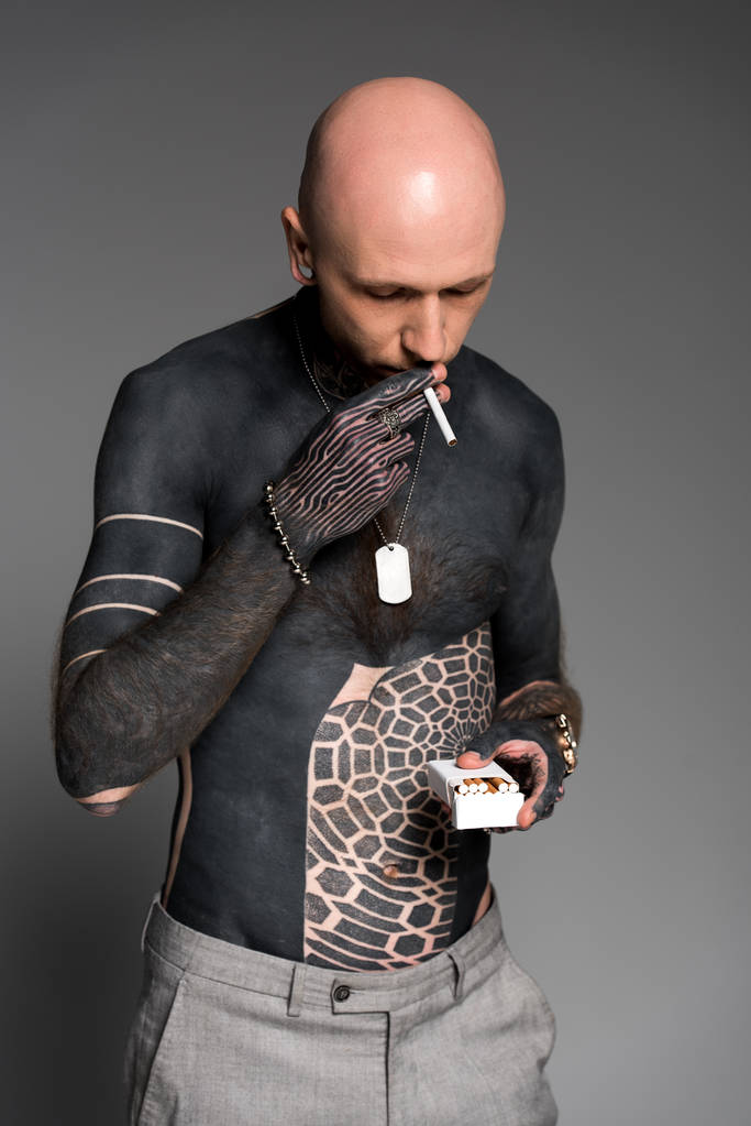 φαλακρός shirtless τατουάζ άνθρωπος που κρατά το κουτί και το κάπνισμα τσιγάρων που απομονώνονται σε γκρι - Φωτογραφία, εικόνα