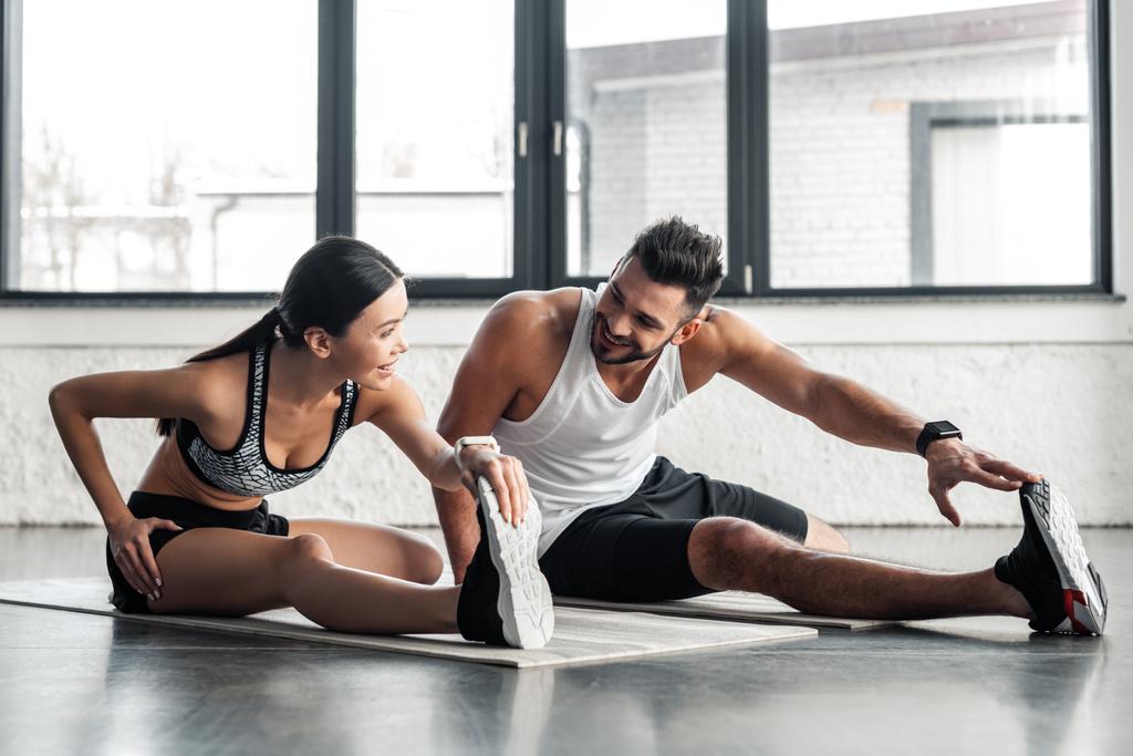 sportieve jonge paar benen strekken en glimlachen elkaar tijdens een training op yoga matten in sportschool - Foto, afbeelding
