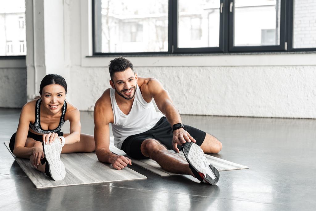 cheerful young sporty couple stretching on yoga mats and smiling at camera in gym - Photo, Image