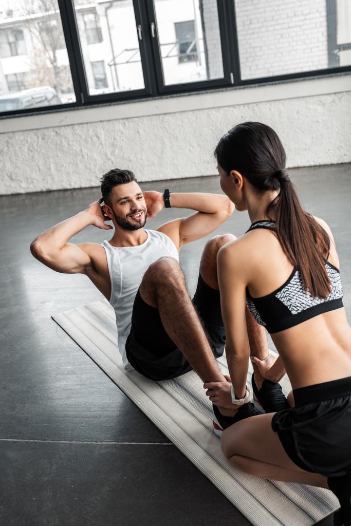 vista de ángulo alto de chica deportiva ayudando a hombre joven feliz haciendo ejercicios abdominales en estera de yoga en el gimnasio
 - Foto, imagen
