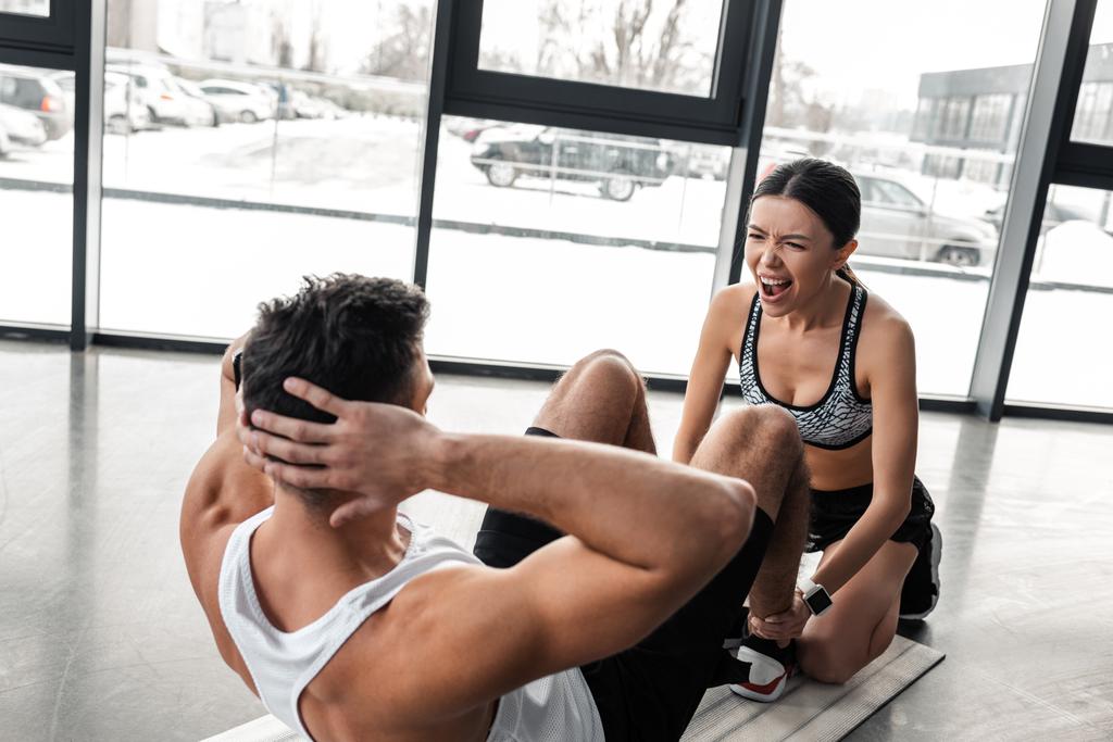 mujer joven deportivo emocional gritando y ayudando a hombre atlético haciendo ejercicios abdominales en el gimnasio
 - Foto, Imagen