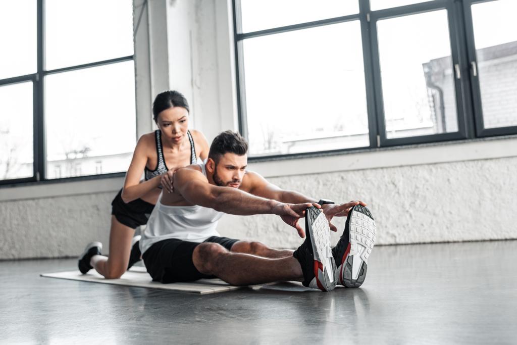 привлекательная спортивная молодая женщина помогает мускулистому спортсмену растягиваться на коврике для йоги в тренажерном зале
  - Фото, изображение