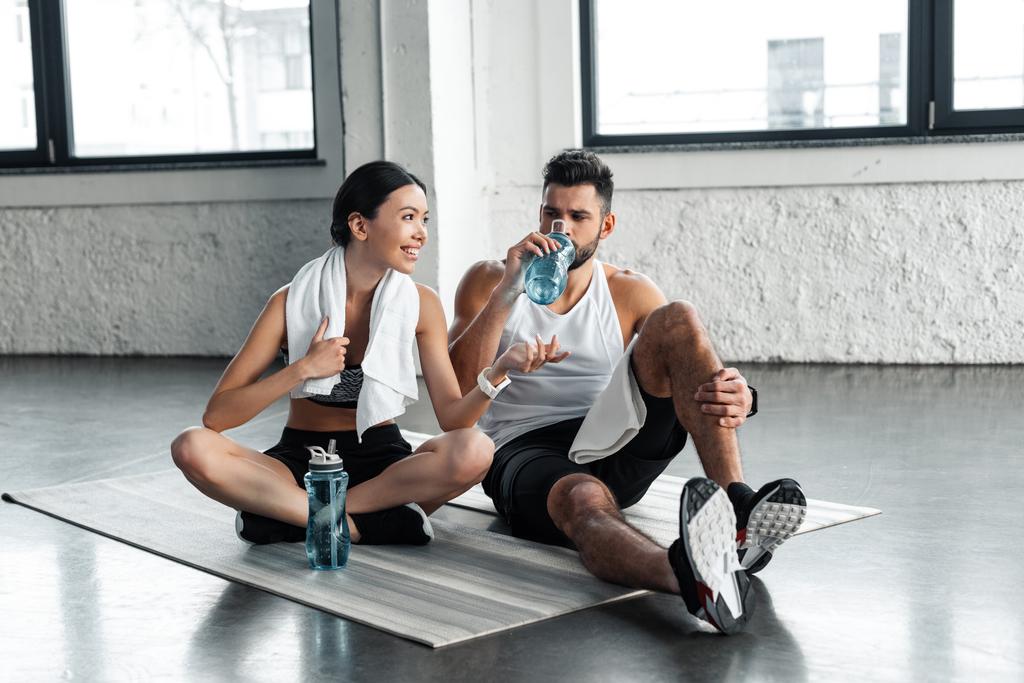 pareja joven atlética bebiendo agua y descansando en colchonetas de yoga después del entrenamiento en el gimnasio
 - Foto, Imagen