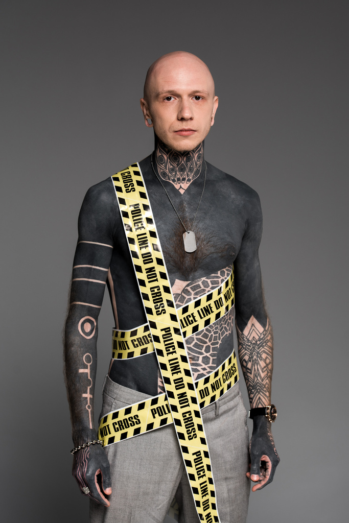 φαλακρός shirtless τατουάζ τον άνθρωπο με αστυνομία γραμμή γύρω από το σώμα που βλέπουν τα φωτογραφικών μηχανών που απομονώνονται σε γκρι - Φωτογραφία, εικόνα