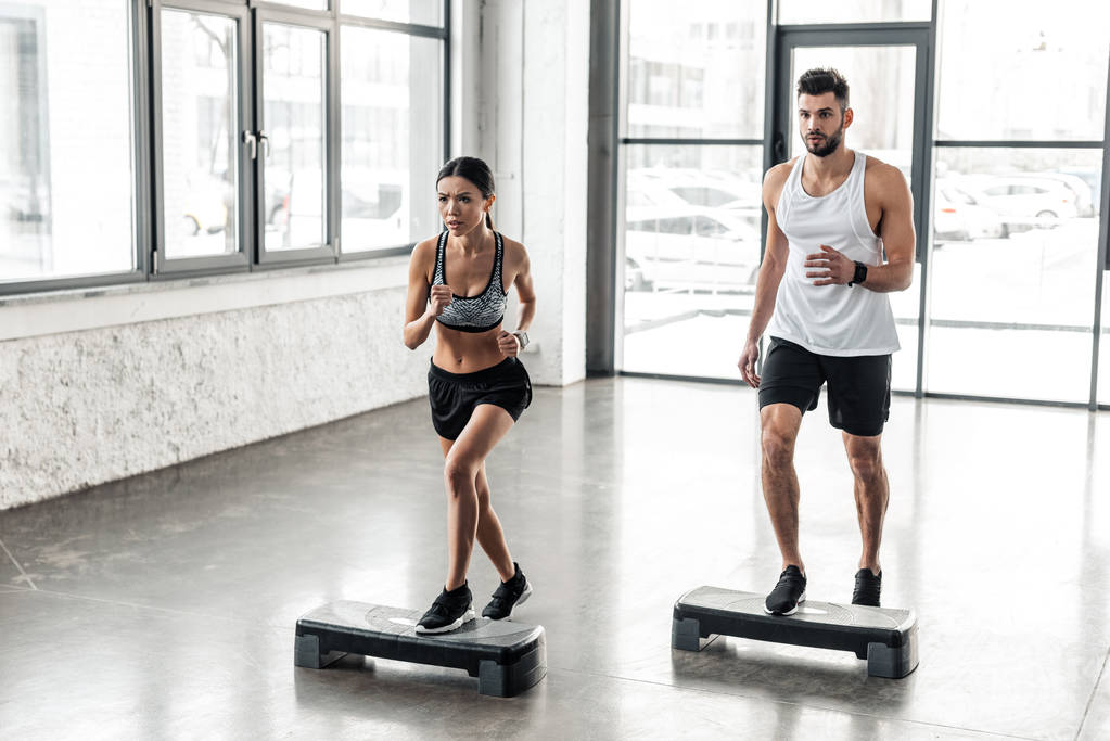 muscular young man and beautiful sporty girl exercising together on step platforms in gym  - Photo, Image