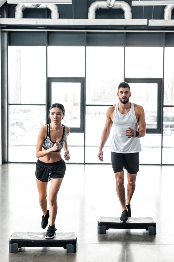 Ganzkörperansicht des sportlichen jungen Paares, das auf Stufenplattformen im Fitnessstudio trainiert  - Foto, Bild