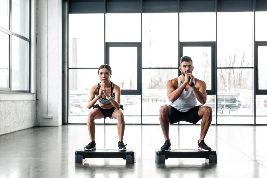 vista completa de la pareja joven atlética en cuclillas en plataformas de paso en el gimnasio
 - Foto, Imagen