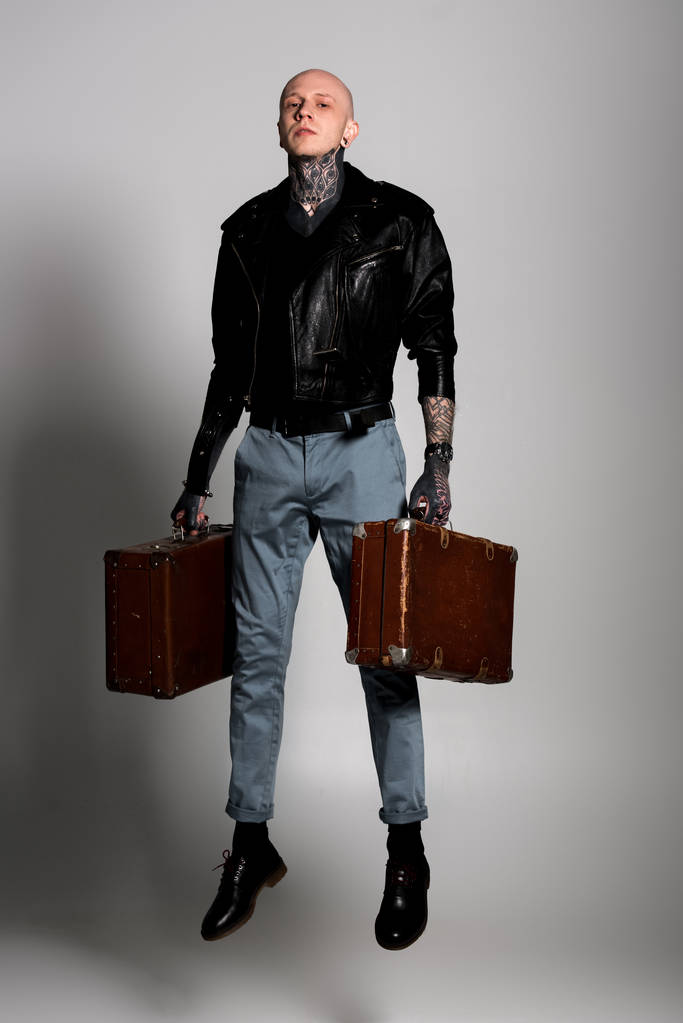 Ganzkörperansicht eines gutaussehenden tätowierten Mannes in Lederjacke, der Koffer hält und auf Grau springt - Foto, Bild