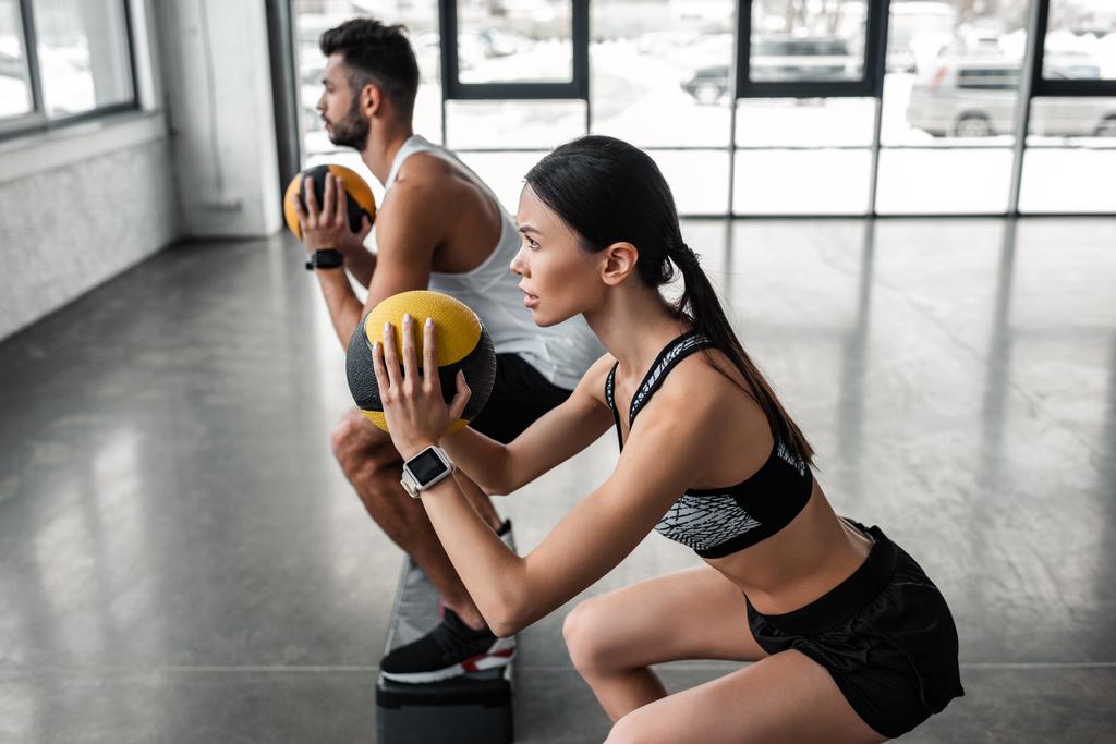 Zijaanzicht van sportieve jonge man en vrouw houden geneeskunde ballen en oefenen met stap platformen in sportschool - Foto, afbeelding