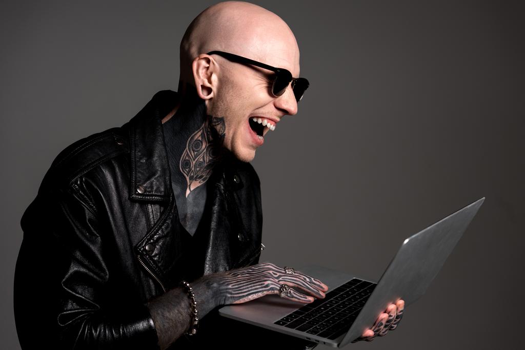 bald tattooed man in leather jacket and sunglasses using laptop and laughing isolated on grey - Photo, Image