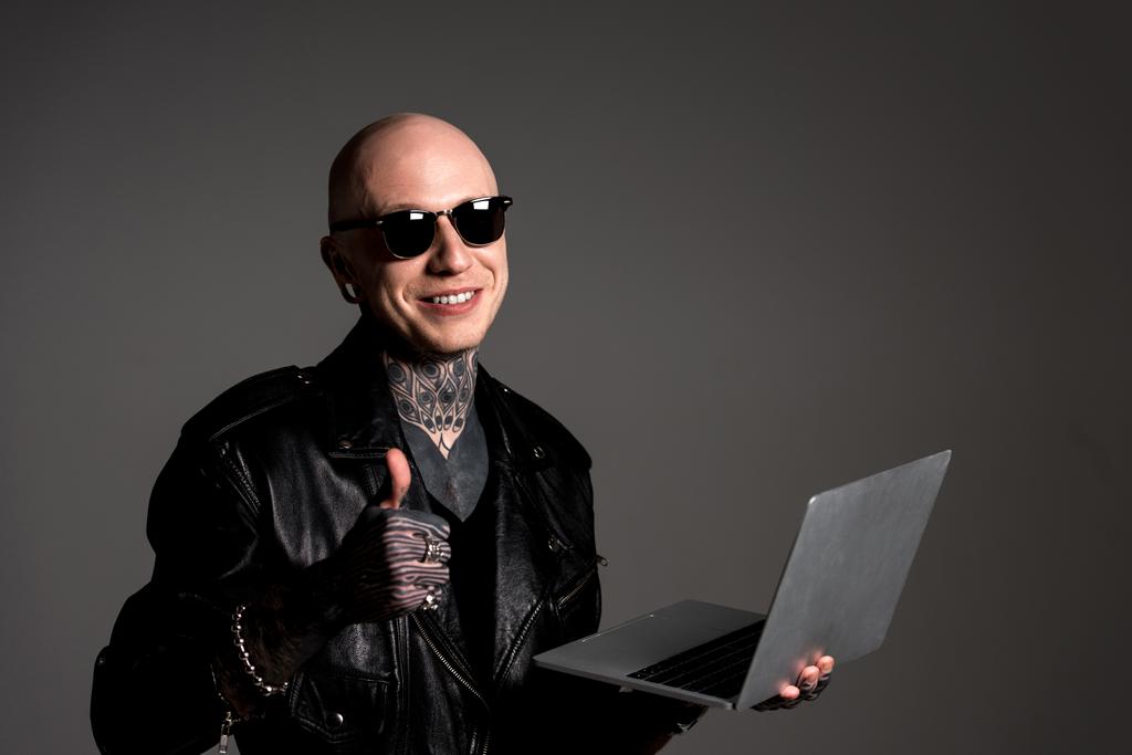 χαμογελώντας τατουάζ τον άνθρωπο σε δερμάτινο μπουφάν και γυαλιά ηλίου, κρατώντας το laptop και δείχνει τον αντίχειρα επάνω απομονώνονται σε γκρι - Φωτογραφία, εικόνα