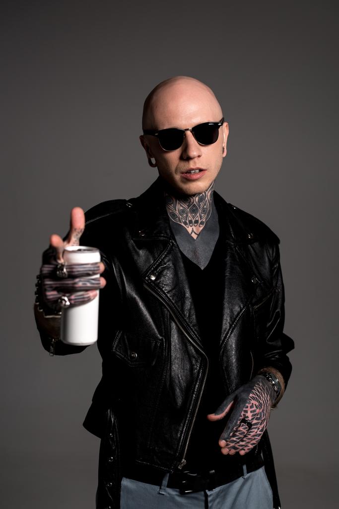 hombre calvo tatuado en chaqueta de cuero y gafas de sol sosteniendo lata de refresco y apuntando a la cámara aislada en gris
 - Foto, imagen