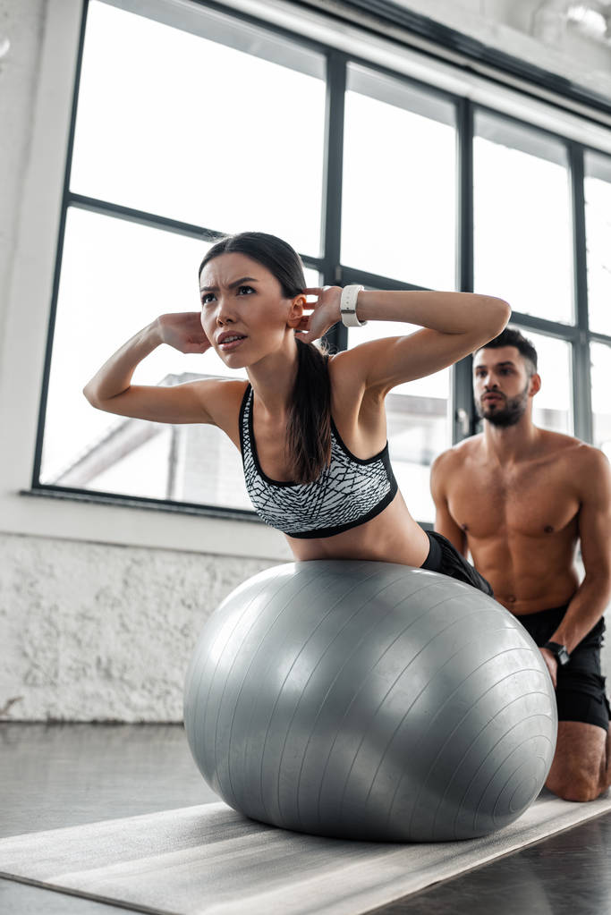nízký úhel pohledu shirtless mužské školitel pomáhá sportovní dívka cvičení na fitness míče v tělocvičně - Fotografie, Obrázek