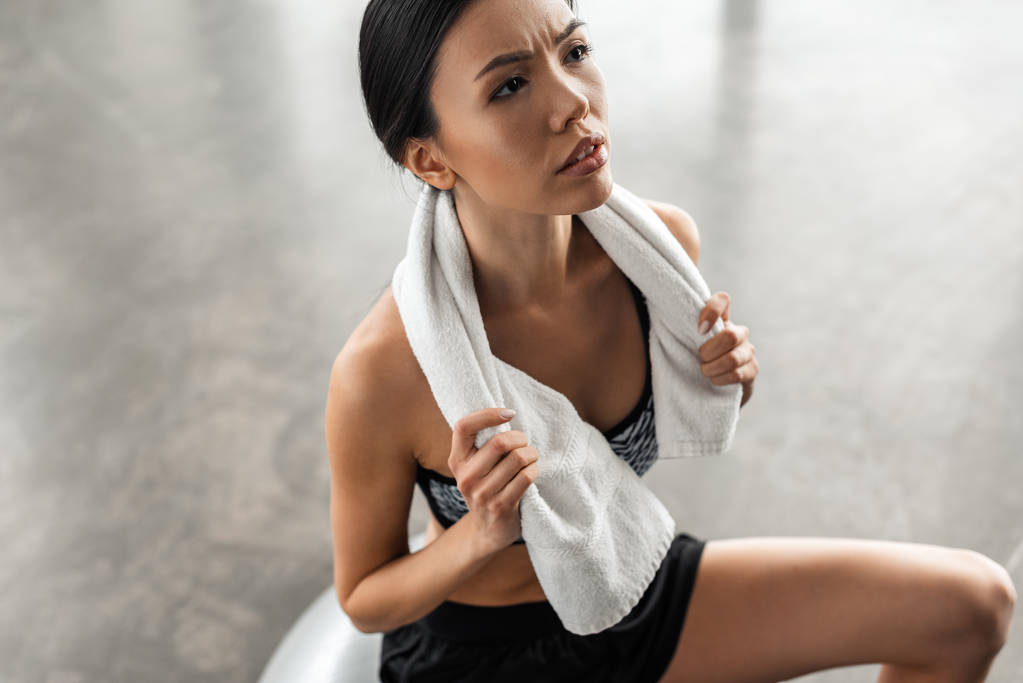 high angle view of serious sporty girl holding towel and looking away while sitting on fit ball in gym - Photo, Image