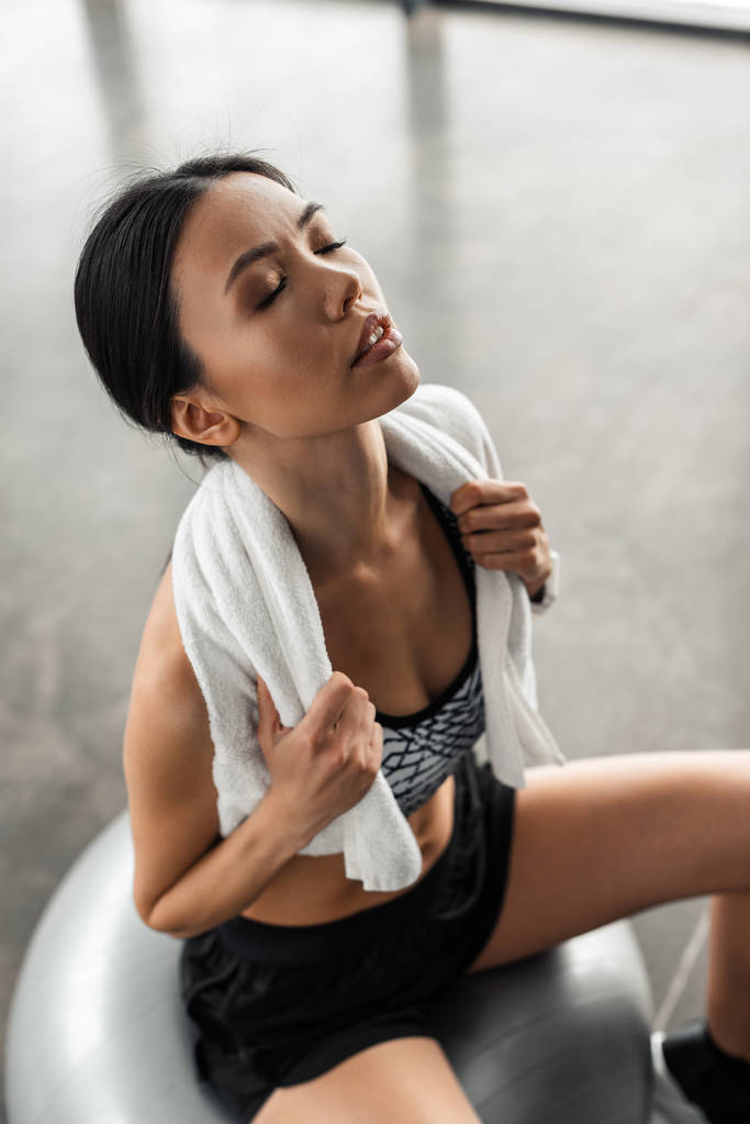 high angle view of exhausted sportswoman with towel on shoulders and closed eyes sitting on fit ball in gym - Photo, Image