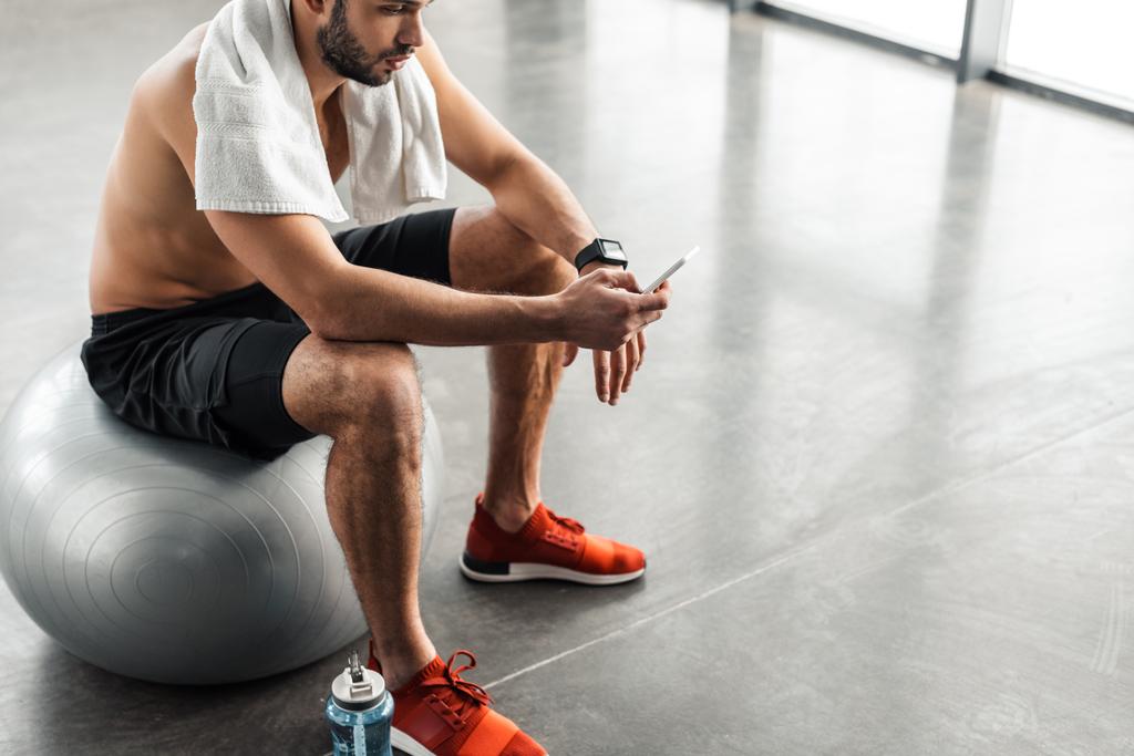 cropped shot of muscular bare-chested man sitting on fit ball and using smartphone in gym - Photo, Image