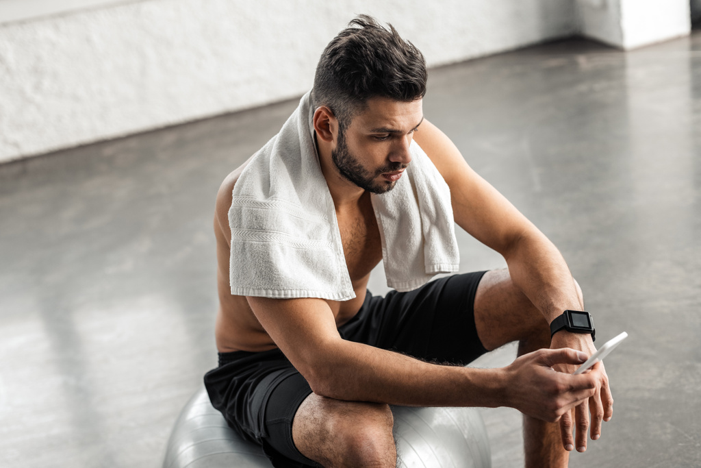 Hochwinkelaufnahme eines muskulösen Mannes mit nacktem Oberkörper, der auf einem fitten Ball sitzt und das Smartphone im Fitnessstudio benutzt - Foto, Bild
