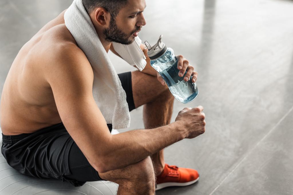 cropped shot of young bare-chested sportsman with towel sitting on fit ball and drinking water in gym - Photo, Image