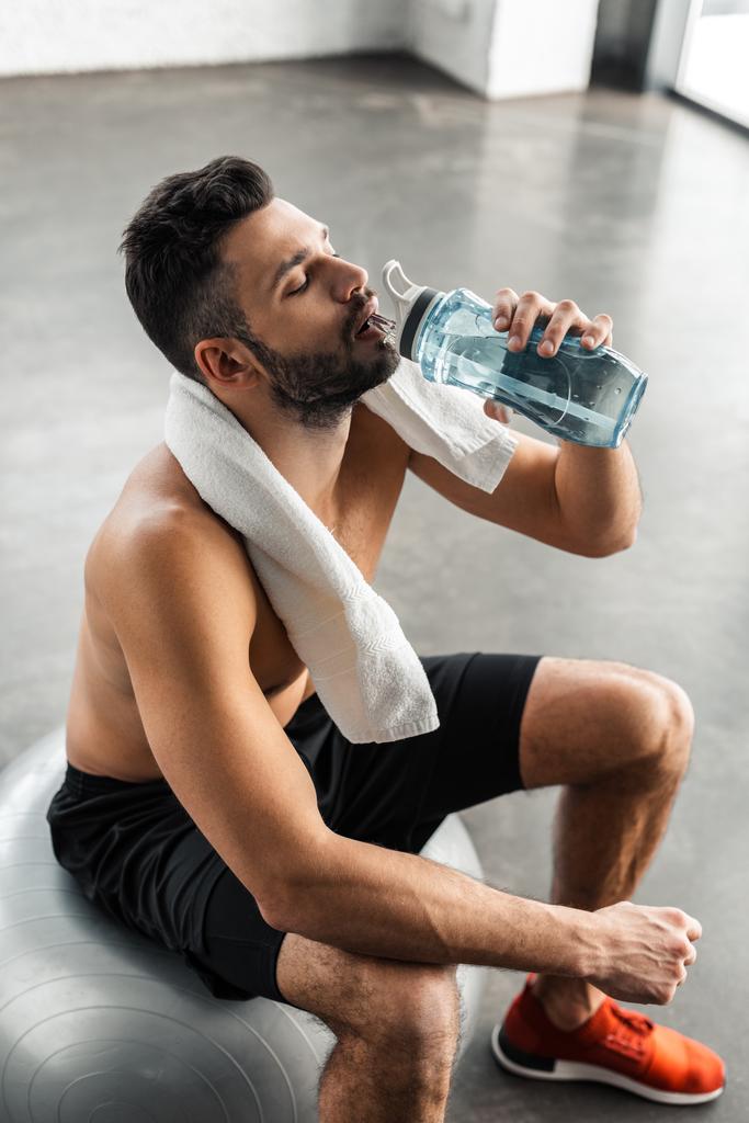 высокий угол зрения молодого спортсмена без рубашки с полотенцем сидя на фитнес мяч и питьевой воды в тренажерном зале
 - Фото, изображение