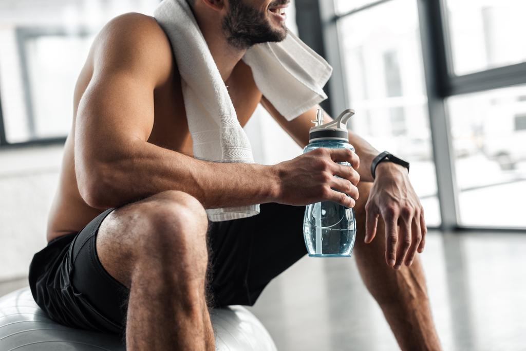 przycięte ujęcie shirtless sportowca z ręcznikiem i butelkę wody, siedząc na fit ball w siłowni - Zdjęcie, obraz