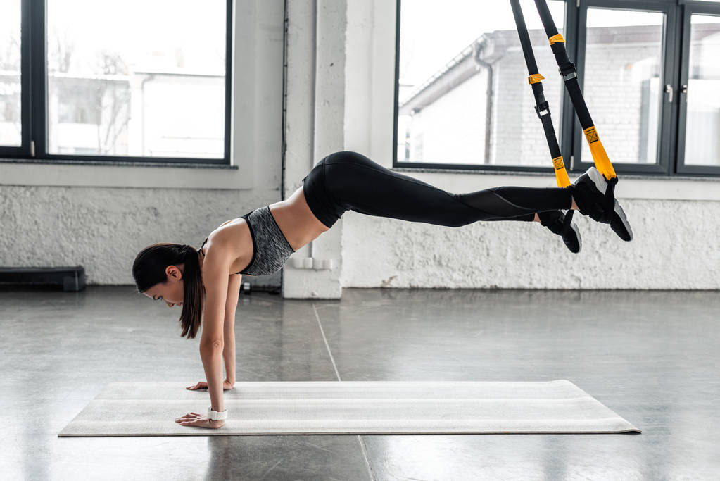 vista laterale di sporty giovane donna che esercita con cinghie di sospensione sul tappetino yoga in palestra
  - Foto, immagini