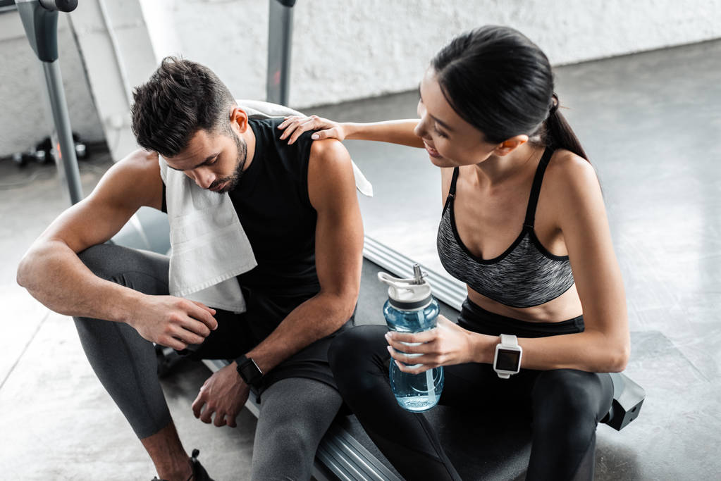 vista de ángulo alto de pareja joven y cansada con toalla y botella de agua descansando juntos en la cinta de correr en el gimnasio
 - Foto, imagen