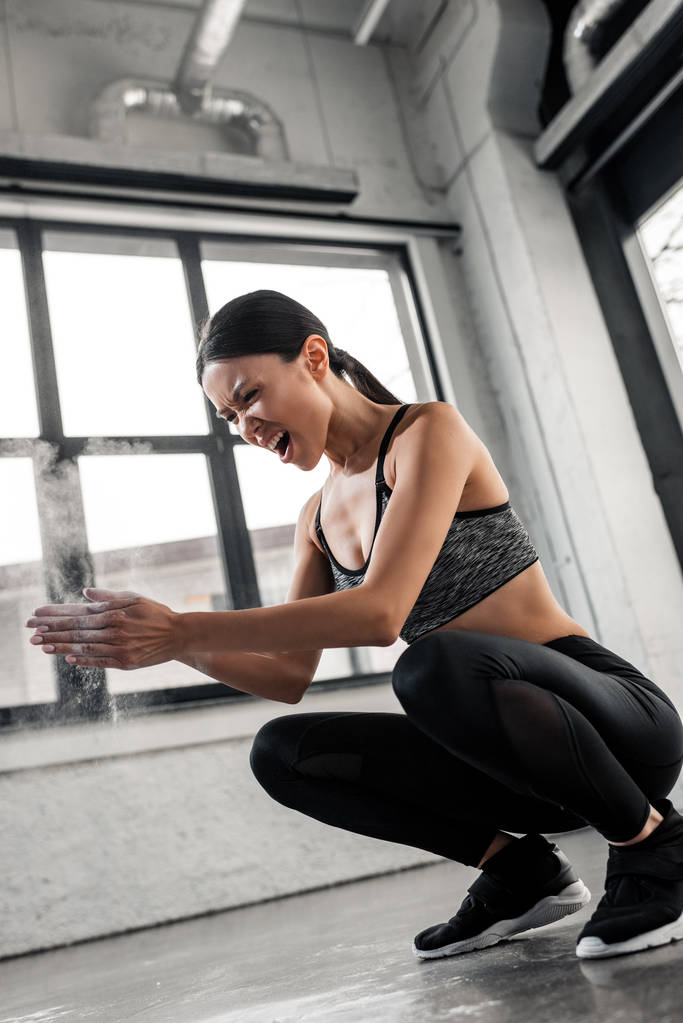 emotional young sportswoman crouching and applying talcum powder on hands in gym - Photo, Image