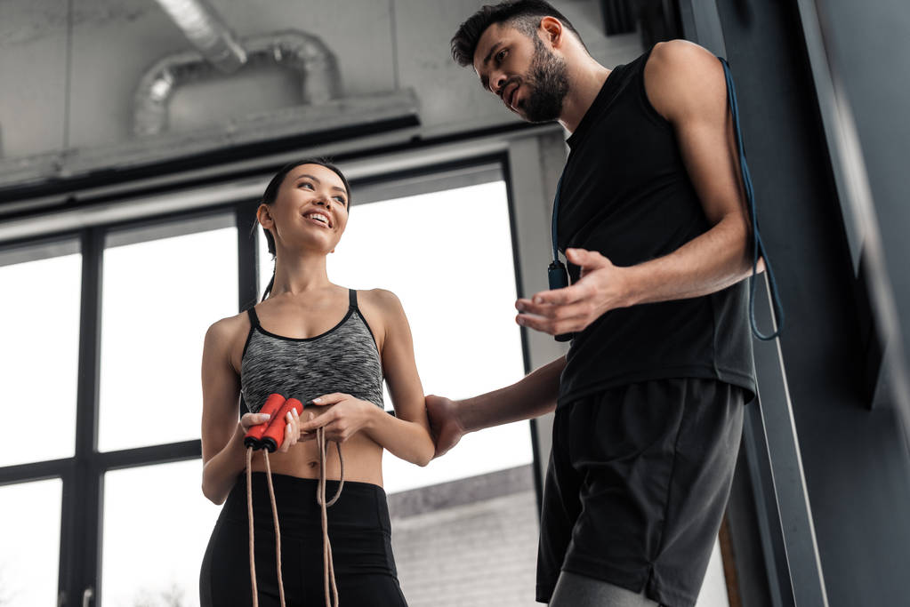 low angle view of smiling sporty couple with skipping ropes standing and talking in gym - Photo, Image