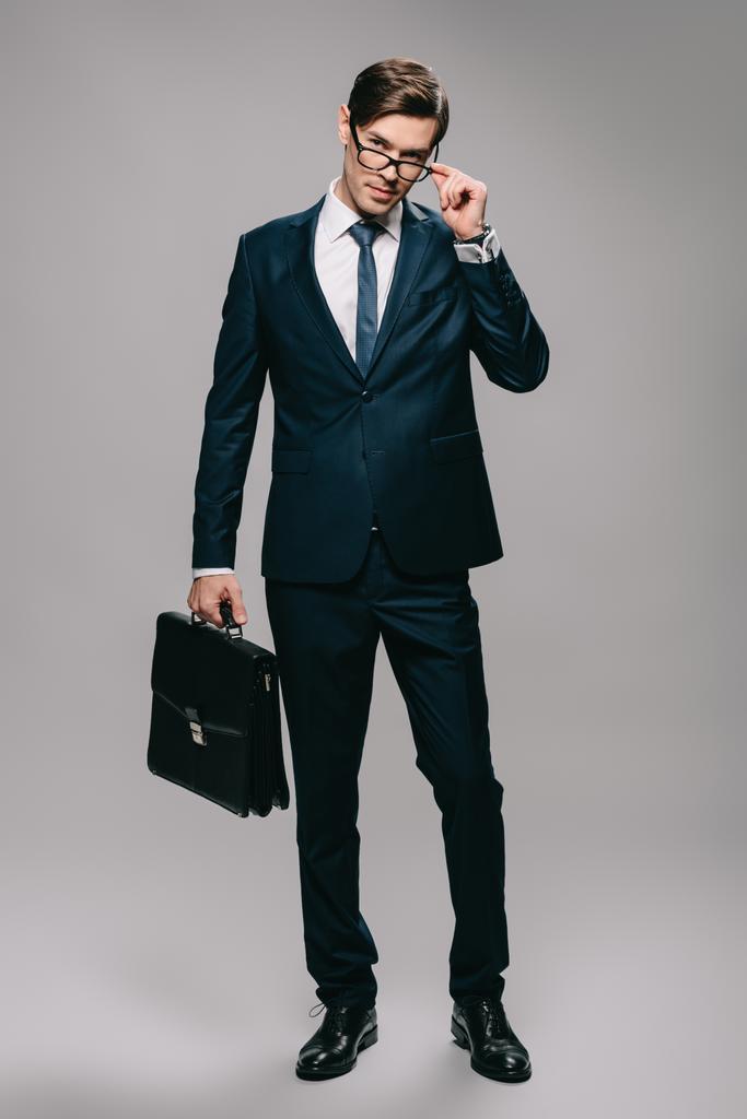confident businessman in suit holding briefcase and touching glasses on grey background  - Photo, Image