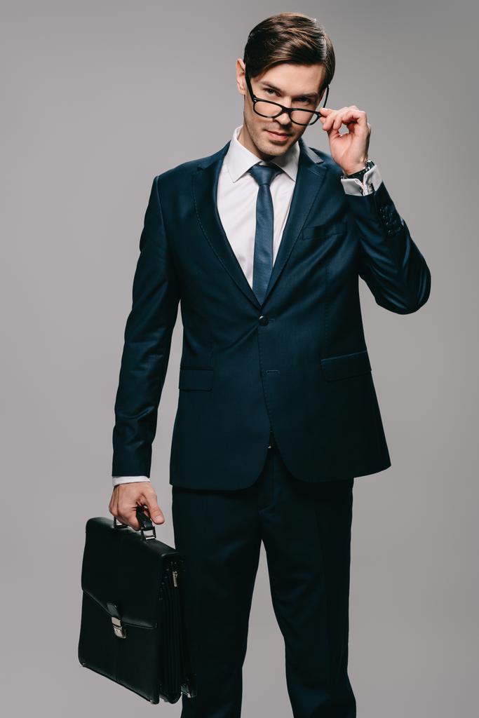 επιχειρηματίας στο κοστούμι κρατώντας χαρτοφύλακα και συγκινητικό γυαλιά σε γκρίζο φόντο  - Φωτογραφία, εικόνα
