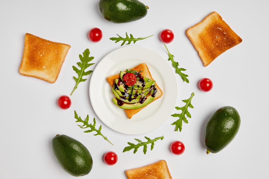 Draufsicht auf Toasts auf Teller mit Avocados, Kirschtomaten und Rucola-Blättern auf grauem Hintergrund - Foto, Bild