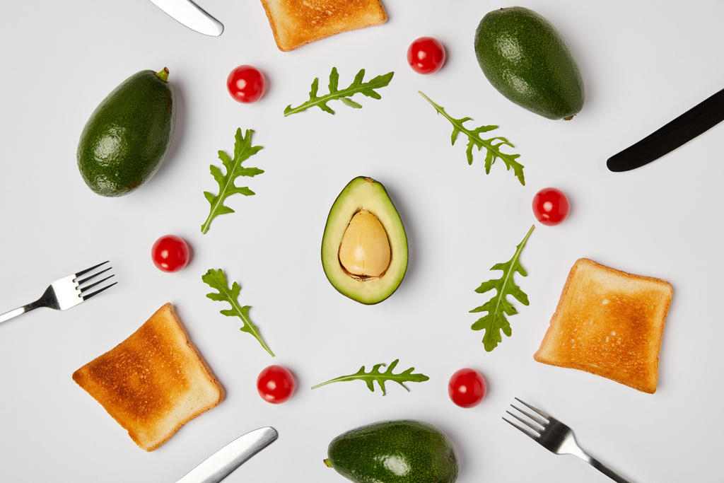 Draufsicht auf Toasts, Avocados, Kirschtomaten und Rucola Blätter, Gabeln und Messer auf grauem Hintergrund - Foto, Bild