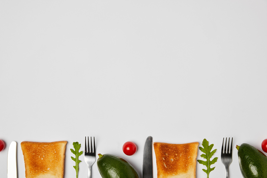 bovenaanzicht van toast, avocado's, kerstomaten, arugulas bladeren, vorken en messen op grijze achtergrond - Foto, afbeelding