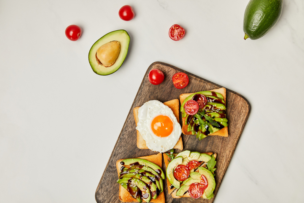 houten snijplank met toast, scrambled ei, avocado's en cherry tomaten op marmeren oppervlak (bovenaanzicht) - Foto, afbeelding