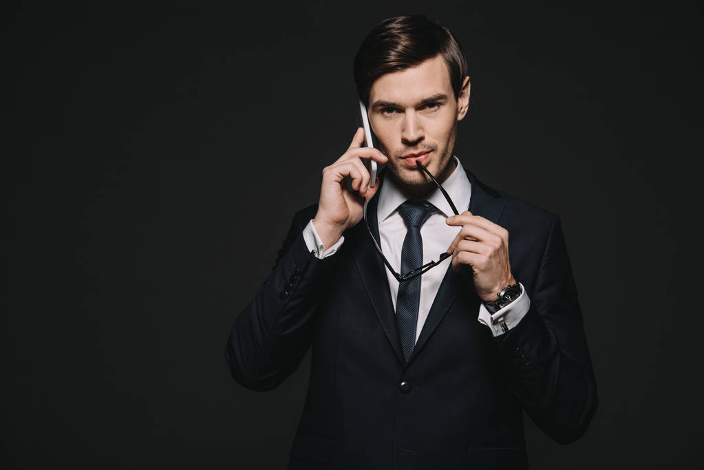 handsome businessman talking on smartphone and holding glasses naer lips isolated on black - Photo, Image