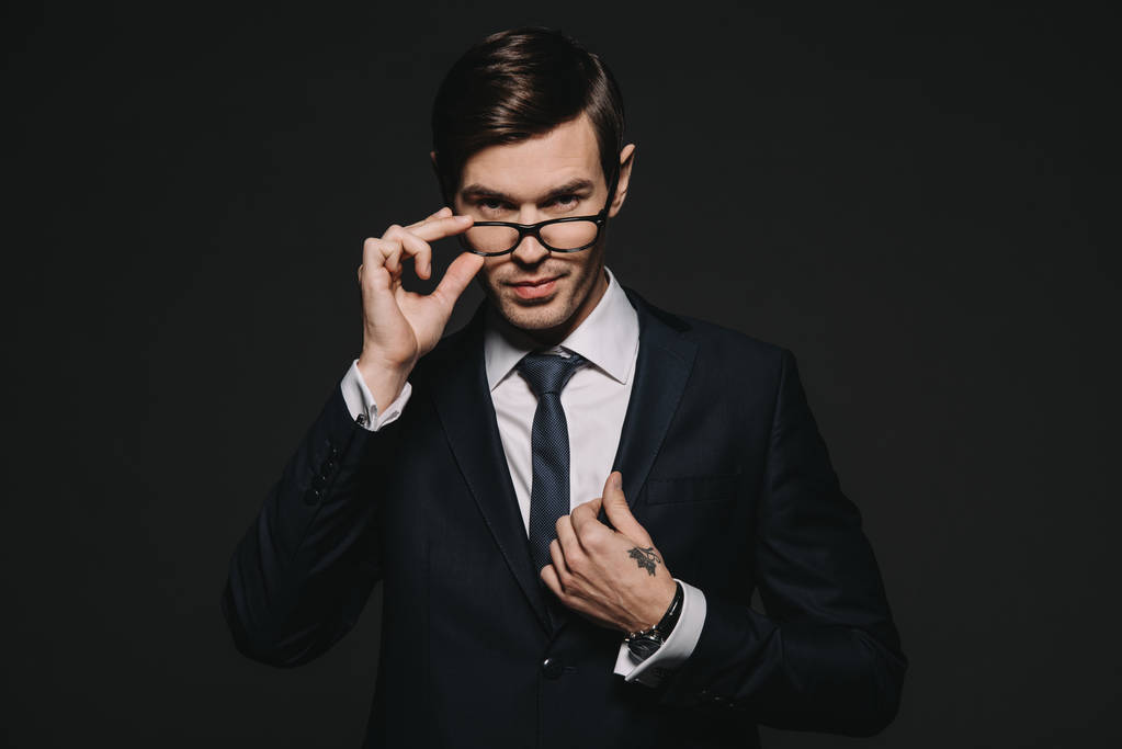 bonito empresário tocando óculos enquanto de pé no terno isolado no preto
 - Foto, Imagem