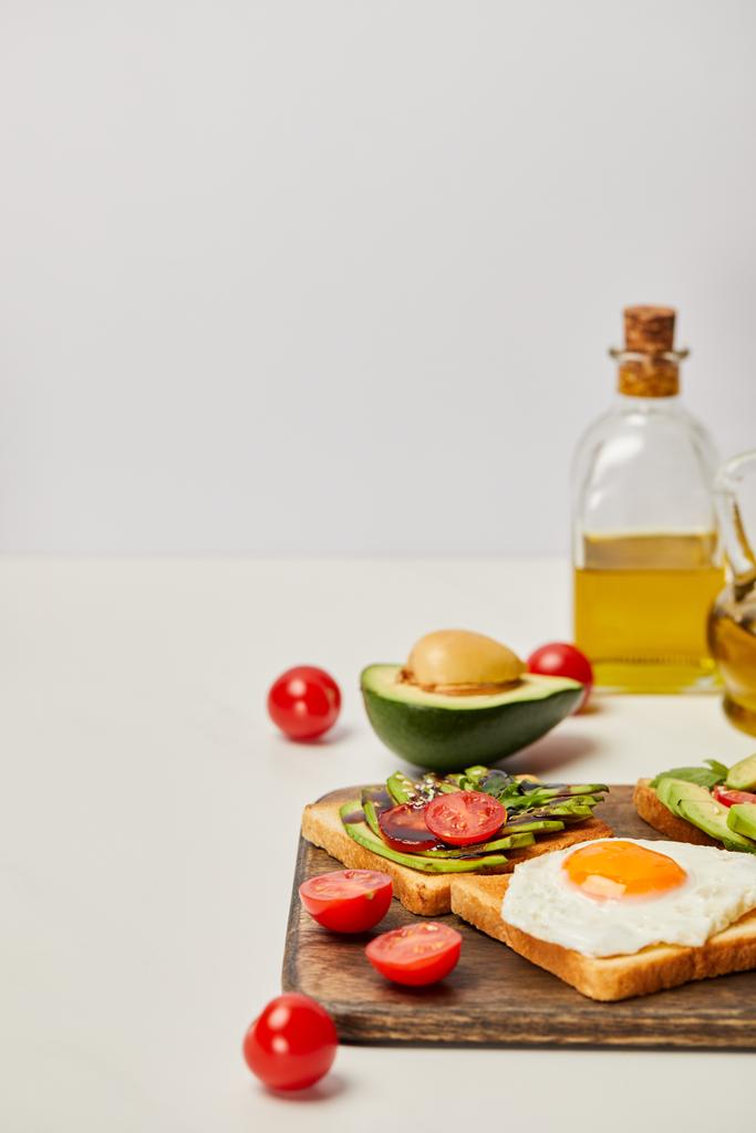 selective focus of wooden cutting board with toasts, scrambled egg, cherry tomatoes, avocados and oil bottles on grey background - Photo, Image