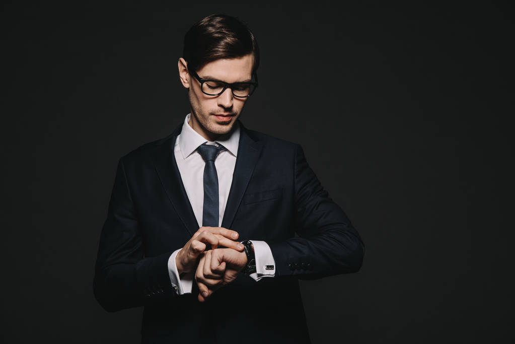 όμορφος επιχειρηματίας ψάχνει στο ρολόι, ενώ στέκεται στο κοστούμι απομονώνονται σε μαύρο - Φωτογραφία, εικόνα