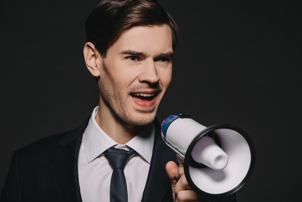 handsome businessman in suit screaming in megaphone isolated on black - Photo, Image