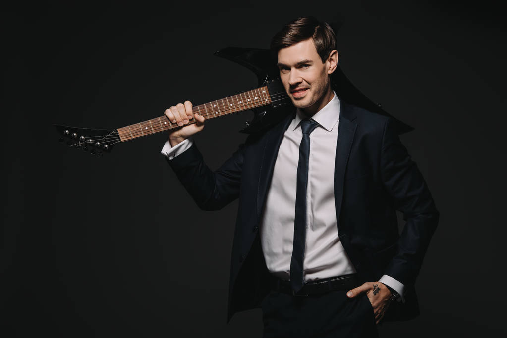 красивый бизнесмен, держащий электро-гитару и стоящий с рукой в кармане, изолированный на черном
 - Фото, изображение