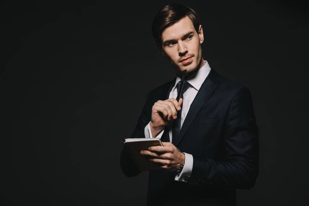 вдумчивый бизнесмен, держащий в руках блокнот и ручку, изолированные от черного
 - Фото, изображение