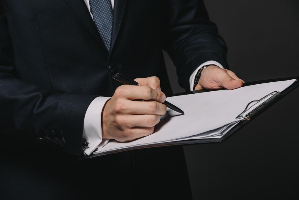 cropped view of businessman writing on paper while holding clipboard isolated on black - Photo, Image