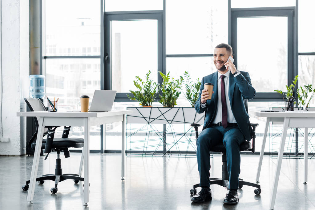 allegro uomo d'affari in possesso di tazza di carta mentre parla su smartphone in ufficio moderno
  - Foto, immagini