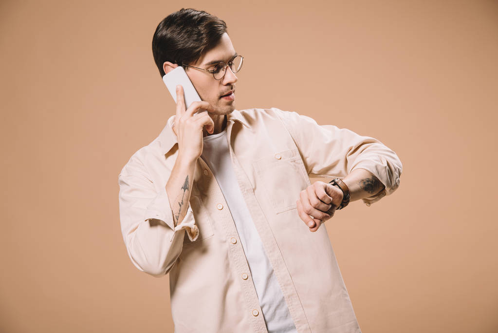 homem bonito em óculos olhando para assistir enquanto fala no smartphone isolado no bege
  - Foto, Imagem