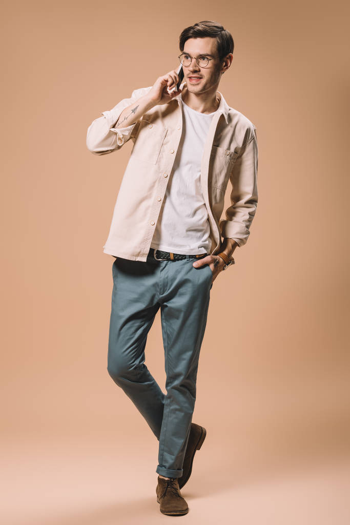 hombre feliz en gafas hablando en el teléfono inteligente mientras está de pie con la mano en el bolsillo sobre fondo beige
 - Foto, Imagen