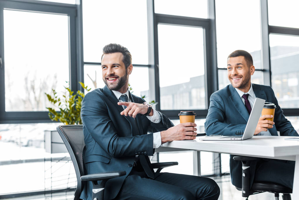 fröhlicher Geschäftsmann zeigt mit dem Finger und hält Pappbecher neben Mitarbeiter in modernem Büro  - Foto, Bild